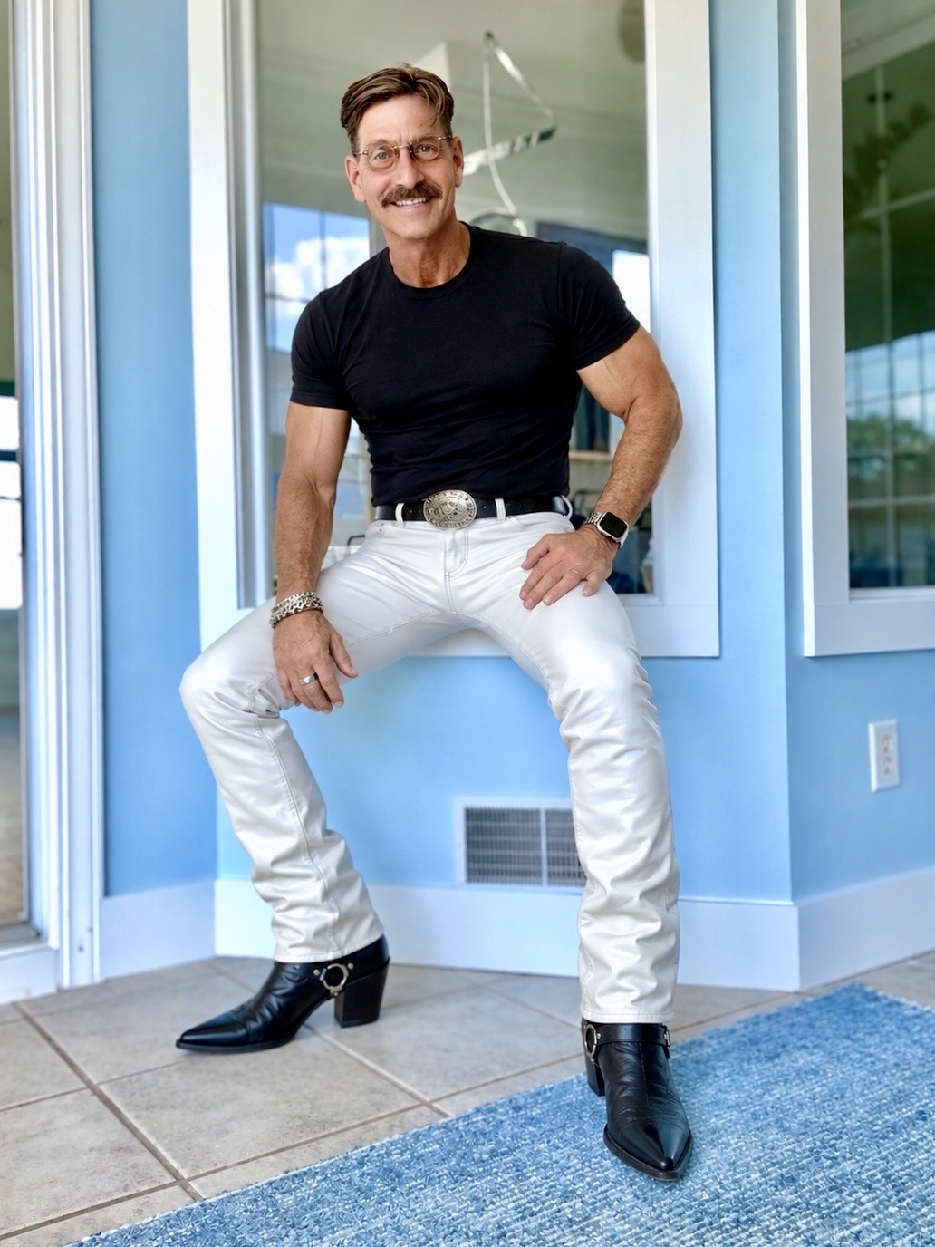 Handsome mature gentleman with thick dark moustache, smiling, sitting on a ledge wearing fitted light denim, black cowboy boots and a black shirt